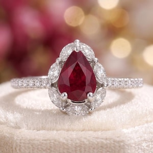 May include: A pear-shaped ruby ring with a halo of marquise-cut diamonds. The ring's band is encrusted with small, round diamonds. The ruby is a deep red color, and the diamonds sparkle against the white metal setting.