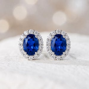 May include: A pair of stud earrings featuring oval, deep blue gemstones surrounded by a halo of smaller, clear stones. The earrings are set in a silver-colored metal, creating a classic and elegant design. The background is blurred, highlighting the jewelry.