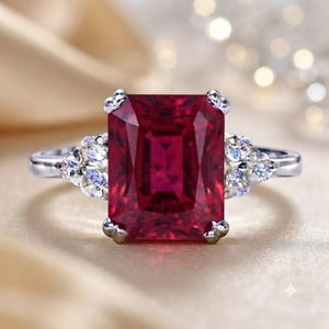 Puede incluir: Un anillo de tono plateado con una gran piedra preciosa rectangular de color rojo intenso como pieza central. La banda está acentuada a ambos lados por un grupo de tres pequeñas piedras claras de corte redondo en engaste de garras.