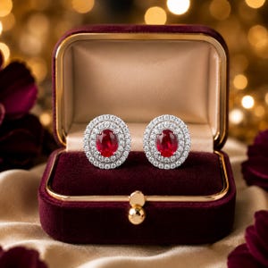 May include: A pair of oval-cut red ruby stud earrings, each surrounded by two concentric halos of small white round-cut diamonds, presented inside a deep burgundy velvet jewelry box with gold-tone metal trim and a secure clasp closure.