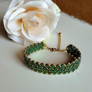 Peut inclure: Un bracelet vert et doré avec un motif tissé. Le bracelet est composé de petites perles vertes rectangulaires et d'éléments dorés. Le fermoir et la chaîne sont également dorés. Une rose blanche est en arrière-plan.
