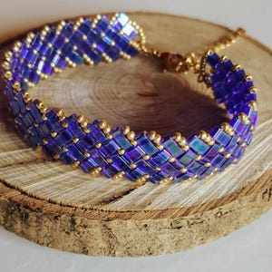 Op de afbeelding: Een handgemaakte armband met een geweven patroon van iriserende blauwe kralen en goudkleurige accenten. De armband wordt gepresenteerd op een houten oppervlak, dat het ingewikkelde ontwerp laat zien. De lengte van de armband is verstelbaar.