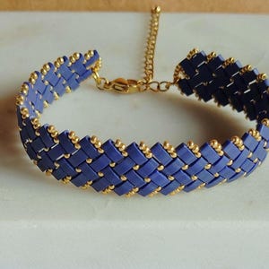 Op de afbeelding: Een geweven armband met rechthoekige blauwe kralen in een diagonaal patroon, geaccentueerd met kleine goudkleurige kralen langs de randen. De armband is voorzien van een goudkleurige metalen verlengketting en een karabijnsluiting.