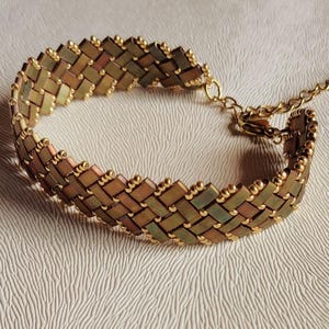 Puede incluir: Una pulsera tejida con cuentas rectangulares metálicas en tonos bronce y verde dispuestas en un patrón diagonal, acentuada con pequeñas cuentas doradas en los bordes y una cadena de extensión de metal dorado con cierre de mosquetón.