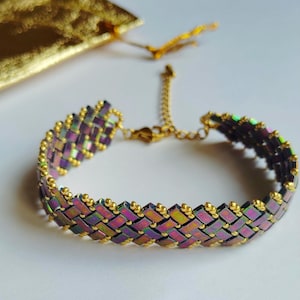 Peut inclure: Bracelet tissé avec des perles rectangulaires irisées aux tons de violet, vert et or. Le bracelet est orné de métal doré et possède un fermoir à chaîne réglable.
