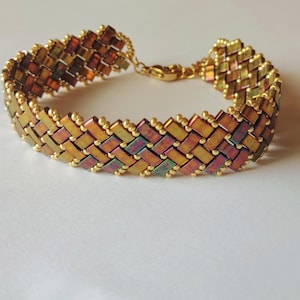 Peut inclure: Un bracelet doré avec un motif tissé. Le bracelet présente des perles rectangulaires dans des tons or, bronze et irisés. Le fermoir et les éléments de connexion sont également dorés. Le bijou est présenté sur une surface blanche.