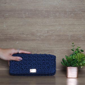 Pode incluir: Uma clutch de crochê azul marinho com formato retangular e uma pequena etiqueta retangular. A clutch é segurada por uma mão com um anel. Uma pequena planta em vaso está à direita da clutch.