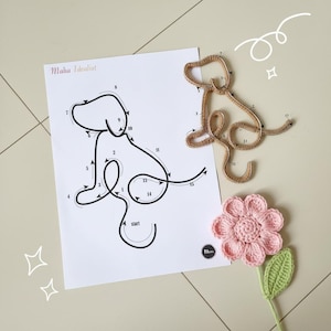 May include: A dog-shaped wire sculpture and a crochet flower with a green stem and leaf are displayed. A paper with a dog outline and the text "Maha Idealist" is also present. The flower is pink, and the wire dog is brown.