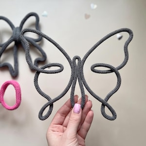 May include: A dark gray butterfly-shaped wall decoration crafted from a continuous length of knitted cord. The design features symmetrical wings with looped edges. In the background, a dark gray bow and a pink script word are visible.