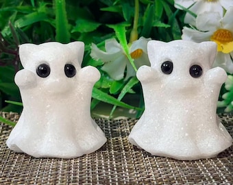 May include: Two small, white, glittery ghost figurines with smooth, rounded shapes and large, glossy black eyes. Each ghost has a wide, flared base and two small, raised arms extending outward, creating a simple, uniform, and symmetrical appearance.