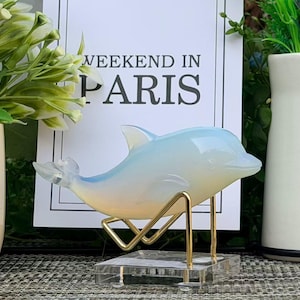May include: An opalescent dolphin ornament in shades of blue and yellow, presented on a gold-coloured metal stand. The dolphin is positioned in front of a sign that reads "WEEKEND IN PARIS". The scene includes vases and greenery.