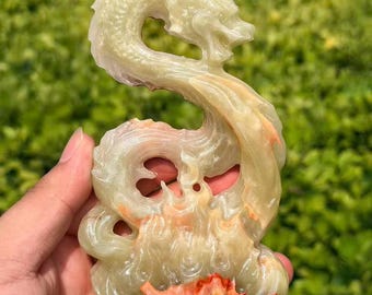 May include: A carved stone sculpture depicting a serpentine dragon figure. The piece features translucent pale green and cream tones with distinct patches of vibrant orange and reddish-brown coloration at the base, showcasing intricate detailing.