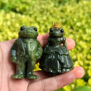 Peut inclure: Deux petites figurines de grenouilles sculptées dans une pierre verte. Une grenouille porte un costume avec une cravate, tandis que l'autre porte une robe à volants et une petite couronne dorée sur la tête. Surfaces polies et yeux ronds.