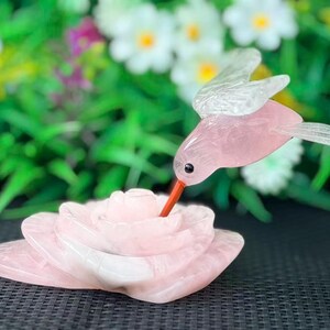 May include: A carved rose quartz hummingbird figurine hovering over a carved rose quartz flower. The hummingbird has translucent wings and a thin orange beak pointed toward the center of the layered pink stone petals of the flower.