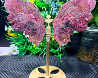 Puede incluir: Una escultura decorativa de mariposa con alas estampadas en rosa y negro. La mariposa está montada sobre un soporte dorado con una base circular. Las alas tienen una apariencia texturizada, y la escultura está sobre un fondo de vegetación.