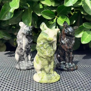 May include: Three carved stone cat figurines in shades of gray, green, and brown. The cats are in a seated position with upright ears and detailed fur textures. The background features green foliage.