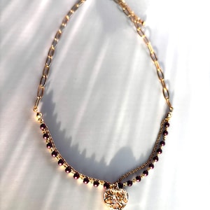 Könnte beinhalten: Goldfarbene Halskette mit zweireihigem Design. Der obere Strang hat kleine, dunkelviolette Perlen, während der untere Strang eine zarte Kette aufweist. Ein herzförmiger Anhänger mit aufwendigen Ausschnitten hängt in der Mitte.