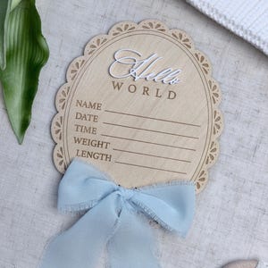 May include: Wooden baby announcement sign with the words "Hello World" in white script. The sign has space to write the baby's name, date, time, weight, and length. A light blue ribbon is tied at the bottom.