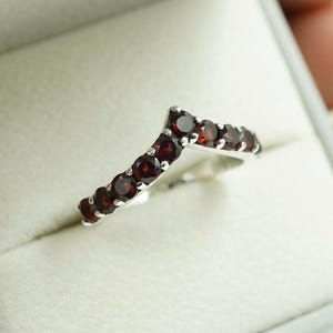 Può includere: Un anello d'argento con un design a forma di V, incastonato con piccole pietre preziose rotonde, rosso scuro. L'anello è presentato in un cofanetto grigio, evidenziando il suo aspetto elegante e delicato.