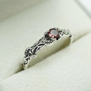 Pode incluir: Um anel de prata com uma pedra preciosa redonda, vermelho escuro. O anel apresenta um design detalhado de videira e folhas. O anel é apresentado numa caixa branca.