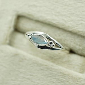 May include: A silver ring featuring a light blue, marquise-cut gemstone. The band has a flowing design with small decorative elements. The ring is presented against a neutral background. The gemstone is approximately 1.5 cm in length.