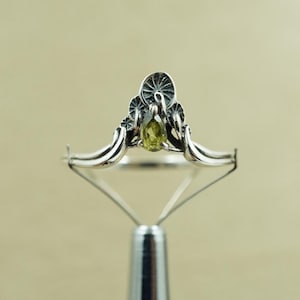 Op de afbeelding: Zilveren ring met een olijfgroene edelsteen in het midden. De ring heeft een decoratieve bovenkant met gestileerde bloemen- of zonnestraalontwerpen en een slanke band.