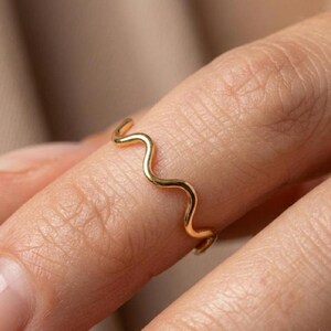 May include: A thin gold ring featuring a continuous wavy or serpentine design. The band has a smooth, polished metallic finish and follows a repeating curved pattern that encircles the finger, creating a delicate and simple aesthetic.
