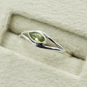 Puede incluir: Un anillo de plata con una piedra preciosa verde claro en forma de pera. La banda del anillo tiene un diseño abierto único, con la piedra preciosa engastada en el centro. El anillo se muestra sobre una superficie beige claro texturizada.