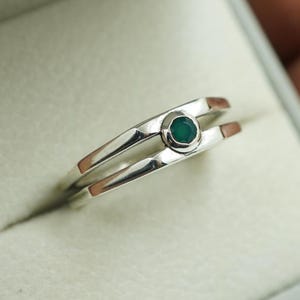 May include: A silver ring with a small, round, emerald-green gemstone set in the center. The ring has a split-shank design, with two parallel bands that meet at the gemstone. The ring is displayed in a gray ring box.