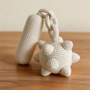 May include: A cream-colored, crocheted toy with a cylindrical handle and a spiky ball shape. The handle is connected to the ball with a series of crocheted rings. The toy is made of a soft, textured yarn and sits on a wooden surface.