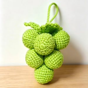 May include: A handmade crochet bunch of grapes in a vibrant green color. The grapes are round and clustered together, with a green stem and leaves. The crochet work is detailed, creating a textured appearance. The grapes are approximately 4 inches tall.