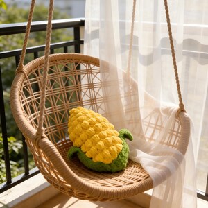 May include: A yellow and green crochet corn plushie with a textured, bumpy kernel design and green husk leaves at the base, resting inside a light brown woven rattan hanging chair suspended by thick ropes on a balcony with sheer curtains.