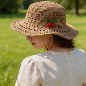 May include: A tan crochet sun hat with a wide brim and decorative cutouts. The hat is adorned with a green leaf and red cherry embellishment. The hat is worn outdoors with a green background.