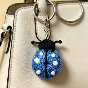 Op de afbeelding: Een gehaakte lieveheersbeestje sleutelhanger met een blauw lichaam met witte stippen en een zwarte kop met voelsprieten, bevestigd aan een zilverkleurige metalen ring. De sleutelhanger hangt aan het handvat van een gladde, gebroken witte tas.
