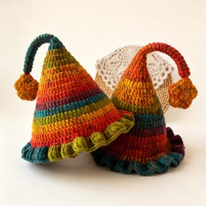 May include: Two handmade, crochet hats in a rainbow of colours. The hats are cone-shaped with a ruffled brim and a curved stem with a star-shaped pom-pom. The colours include red, orange, yellow, green, and blue.