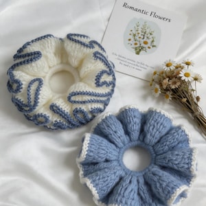 May include: Two circular crochet hair scrunchies. One is white with blue yarn detailing in a swirling pattern. The other is light blue with a white crocheted edge. A small card with the text Romantic Flowers and a small bouquet of dried daisies.