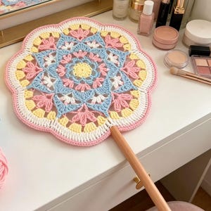 Könnte beinhalten: Ein runder, handgehaltener Häkelfächer mit einem Mandala-inspirierten Muster in Pastelltönen wie Rosa, Hellblau, Gelb und Weiß. Das kreisförmige Textilstück ist an einem glatten, hellen Holzgriff befestigt, der von der Unterkante absteht.