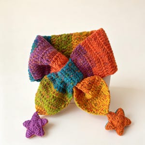 May include: A colorful, handmade crochet scarf with star accents. The scarf features a vibrant mix of orange, purple, green, and blue hues. Two star-shaped embellishments, one purple and one orange, dangle from the ends. The scarf is displayed on a white surface.