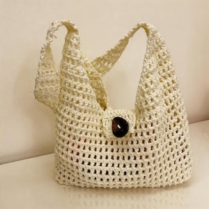 May include: Cream-colored, crocheted tote bag with a large, dark brown button closure. The bag features a woven, open-weave design and two shoulder straps. The bag's interior is partially visible.