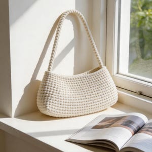 May include: A cream-coloured, hand-crocheted shoulder bag featuring a dense, textured stitch pattern and a single, thick, braided handle. The bag has a structured, rectangular shape with rounded corners and is crafted from a solid, light-coloured yarn.