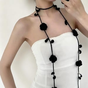 Può includere: Collana nera all'uncinetto con design a girocollo e lungo. Presenta dettagli di fiori e foglie neri. La collana è indossata sopra un top bianco senza spalline. Un accessorio elegante.