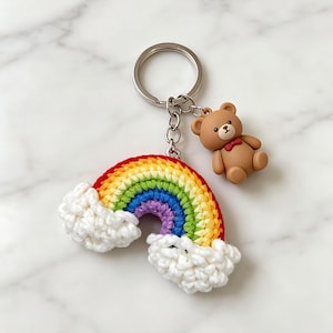Puede incluir: Un llavero con un arcoíris tejido a ganchillo en colores vibrantes y nubes blancas esponjosas. Un pequeño oso de peluche marrón con una pajarita roja está adjunto. El llavero tiene un anillo plateado. El arcoíris es de rojo, naranja, amarillo, verde, azul y morado.