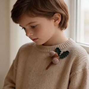 May include: A beige sweater with a crocheted acorn brooch. The brooch features two light brown acorns, green leaves, and a brown stem. The sweater has a ribbed crew neck.
