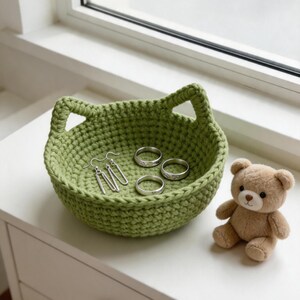 May include: A sage green crocheted basket with two integrated handles shaped like cat ears. Inside the basket are a pair of silver dangle earrings and three silver rings. A small, light brown plush teddy bear sits next to the basket.