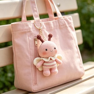 May include: A light pink canvas tote bag with a vertical zippered pocket, featuring a crocheted bee keychain attached to the handle. The bee has a round pink and white striped body, small white wings, brown antennae, and a stitched smiling face.