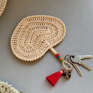 Può includere: Un portachiavi all'uncinetto color crema con un design rotondo e testurizzato a forma di ventaglio. L'impugnatura è avvolta da filo multicolore e include una nappa rosso vivo, un piccolo ciondolo a forma di cuore nero e tre chiavi in metallo.