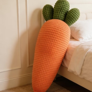 Puede incluir: Un cojín grande con forma de zanahoria que presenta un cuerpo texturizado de ganchillo naranja y tres hojas de ganchillo de color verde oscuro. El cojín tiene una construcción gruesa de hilo con un patrón de puntos visible en la superficie.