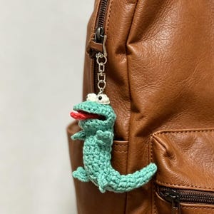 Puede incluir: Un llavero de ganchillo hecho a mano con un caimán verde claro con una lengua roja y ojos blancos. El caimán está sujeto a una cadena plateada y cuelga de una mochila de cuero marrón.