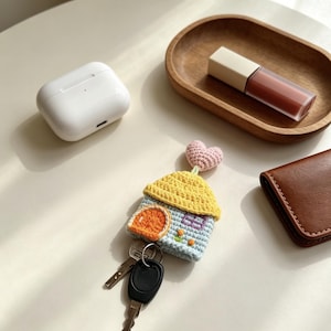 Könnte beinhalten: Eine gehäkelte Schlüsselhülle in Form eines kleinen Hauses mit hellblauer Basis, gelbem Dach, orangefarbener Tür und einem rosa Herz an der Spitze. Das Haus hat ein kleines Fenster und zwei winzige orangefarbene Blumen sowie einen Metallring mit zwei Schlüsseln.