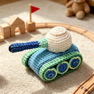 Puede incluir: Un tanque de juguete tejido a ganchillo con cuerpo azul, torreta color crema y un cañón largo azul. El tanque tiene orugas verdes con ruedas circulares azules y verde claro. La construcción utiliza hilo en tonos azul, crema y verde.
